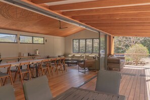 Ein geräumiges Zimmer mit Holzbalkendecke, einem Essbereich mit langem Tisch und Stühlen, einer Couch und einem Fenster mit Blick auf einen Garten.