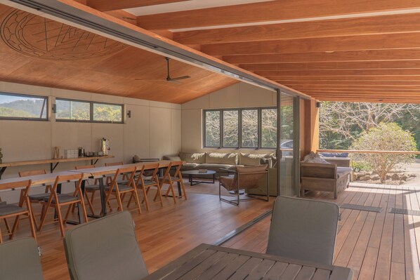 A spacious room with a wooden ceiling, a dining area with a long table and chairs, a sofa, and a window overlooking a garden.