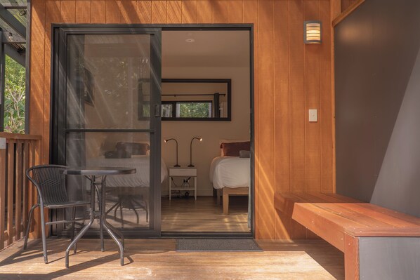 A modern room with a sliding glass door leading to a balcony, a bed, bedside table, and a small desk with a lamp.