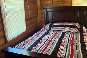 Una cama individual con colcha a rayas y cabecero oscuro en una habitación con paredes de madera y una ventana con persianas.