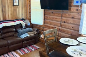 Una habitación con paneles de madera, un sofá de cuero marrón, un televisor montado en la pared y una mesa de comedor con platos.