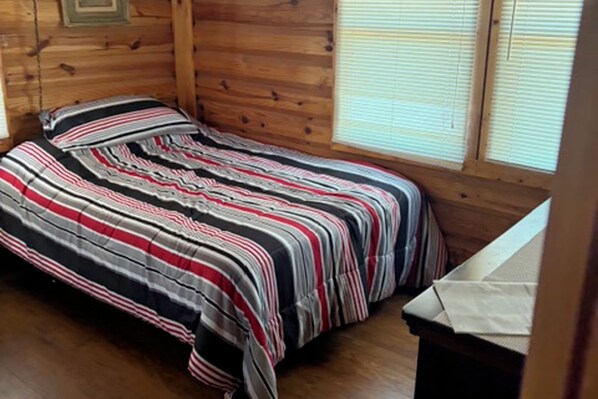 Una habitación de cabaña de madera con una cama, una ventana con persianas y una pequeña mesa.