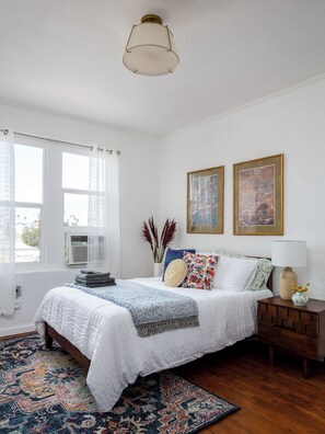 Un dormitorio con una cama, una mesita de noche, una lámpara, una ventana con cortinas y dos cuadros en la pared.