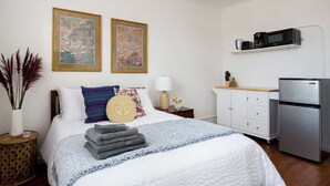 Een verzorgde slaapkamer met een bed, kussens, een nachtkastje met lamp, een klein kastje en een koelkast.