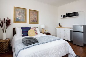 Un dormitorio ordenado con cama, almohadas, una mesita de noche con lámpara, un armario pequeño y un refrigerador.