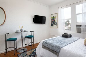 Un dormitorio con una cama, un televisor montado en la pared, una mesita con dos sillas y una ventana con cortinas.