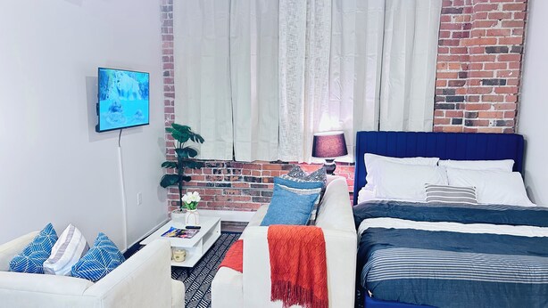 A modern bedroom with a brick wall, a bed with blue bedding, a white sofa with patterned cushions, a small table with a lamp, and a TV on the wall. - Studio in the Heart of
DSM Downtown (Des Moines)