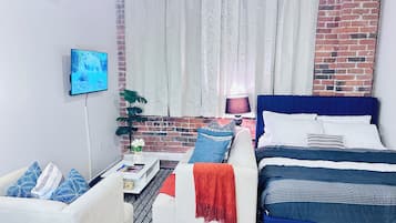 Ein modernes Schlafzimmer mit Backsteinwand, einem Bett mit blauer Bettwäsche, einem weißen Sofa mit gemusterten Kissen, einem kleinen Tisch mit Lampe und einem Fernseher an der Wand.