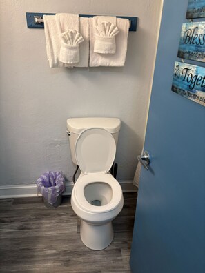 A bathroom with a toilet, a trash bin, and a towel rack with two towels. - Driftwood Studio | Steps to South Myrtle Beach ,Bright, Cozy & Self Check-In (Myrtle Beach)