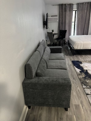 A modern hotel room with a grey sectional sofa, a bed, a small table, and a chair. - Driftwood Studio | Steps to South Myrtle Beach ,Bright, Cozy & Self Check-In (Myrtle Beach)