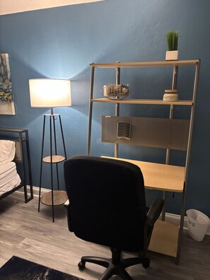 A room with a blue wall, a standing desk, a black office chair, a floor lamp, a shelf with a plant, and a small trash bin. - Driftwood Studio | Steps to South Myrtle Beach ,Bright, Cozy & Self Check-In (Myrtle Beach)