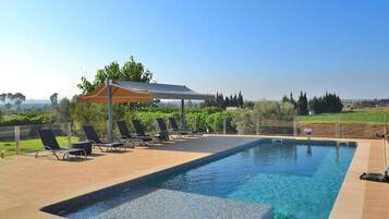 A swimming pool with lounge chairs and a covered area, surrounded by greenery and a clear sky.