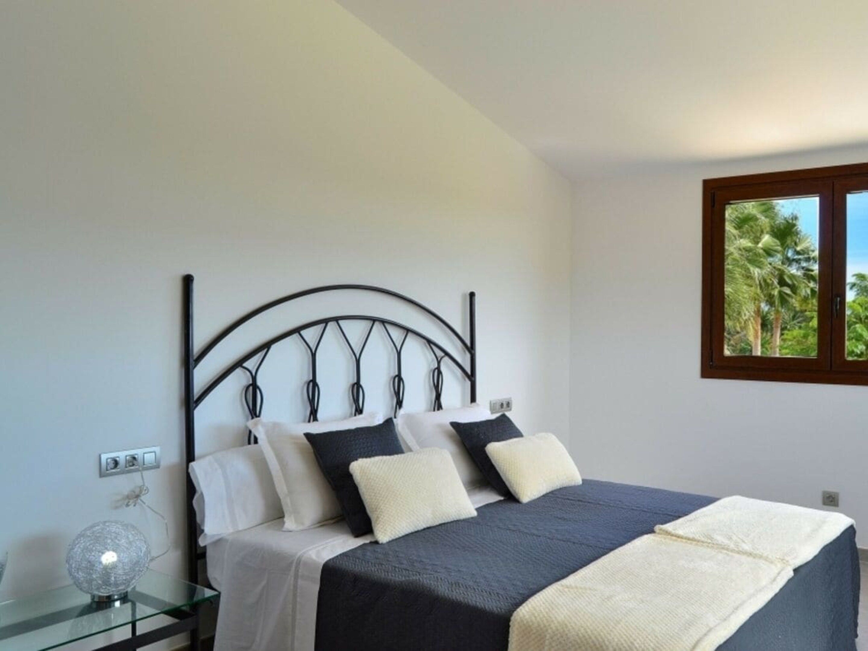 A bedroom with a bed, a bedside table, a lamp, and a window with a view of palm trees.