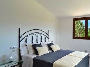 A bedroom with a bed, a bedside table, a lamp, and a window with a view of palm trees. - Cabin in Muro With Swimming Pool (Muro)