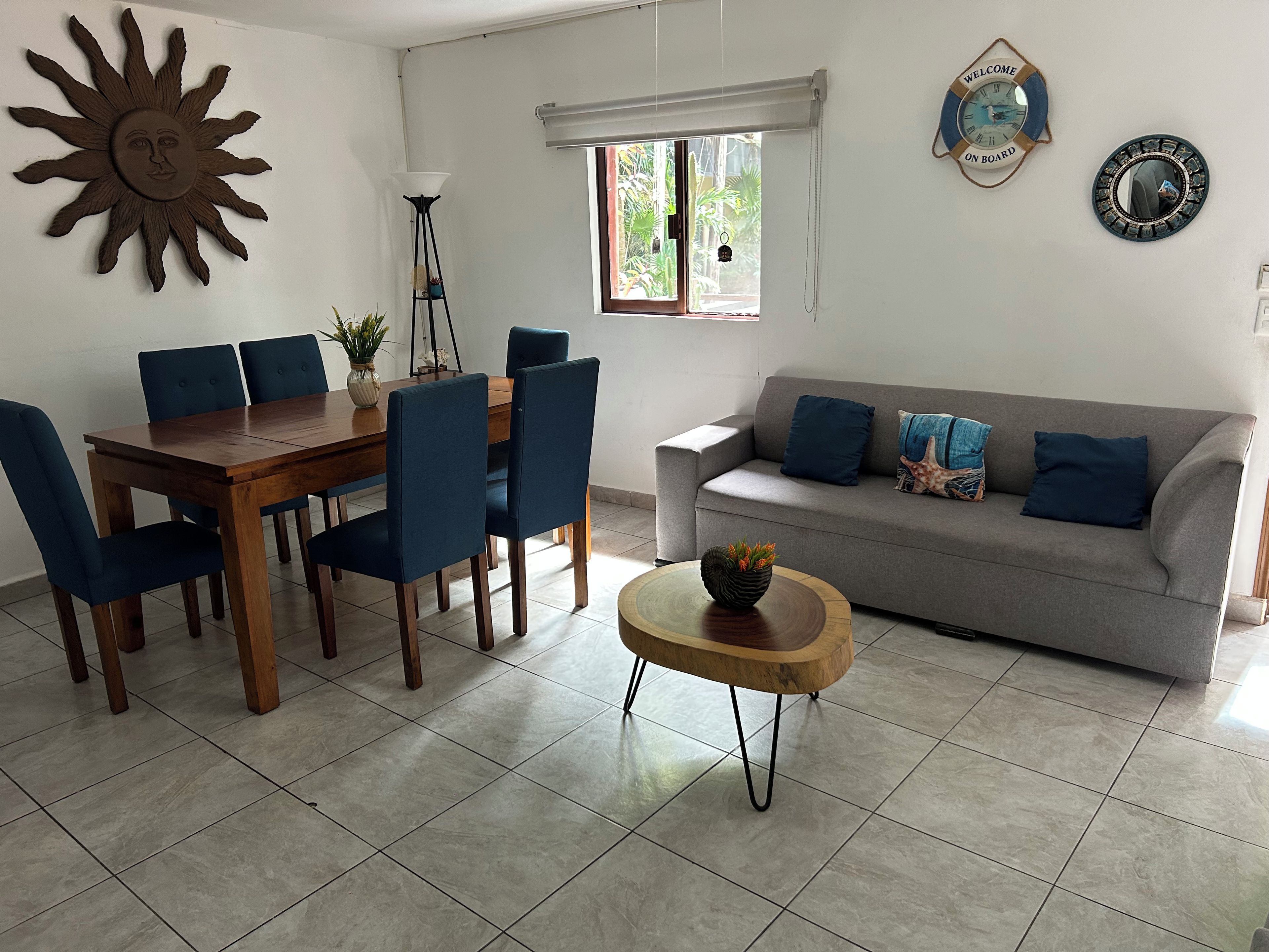 A dining area with a wooden table and blue chairs, a grey sofa with blue cushions, and a decorative sun on the wall.