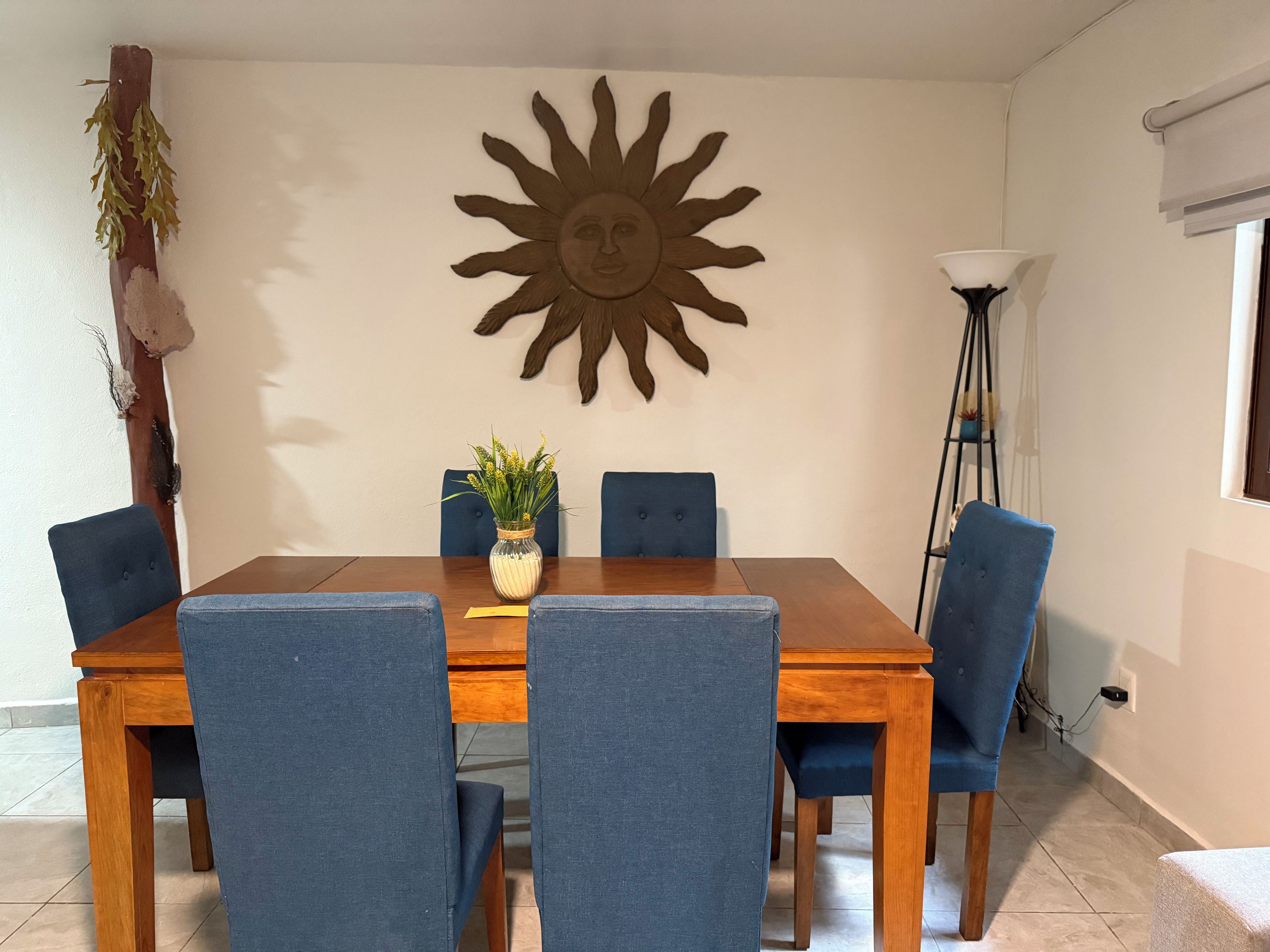 Ein Essbereich mit Holztisch, blau gepolsterten Stühlen, einer Blumenvase und einer Wanddekoration mit Sonnenelementen.