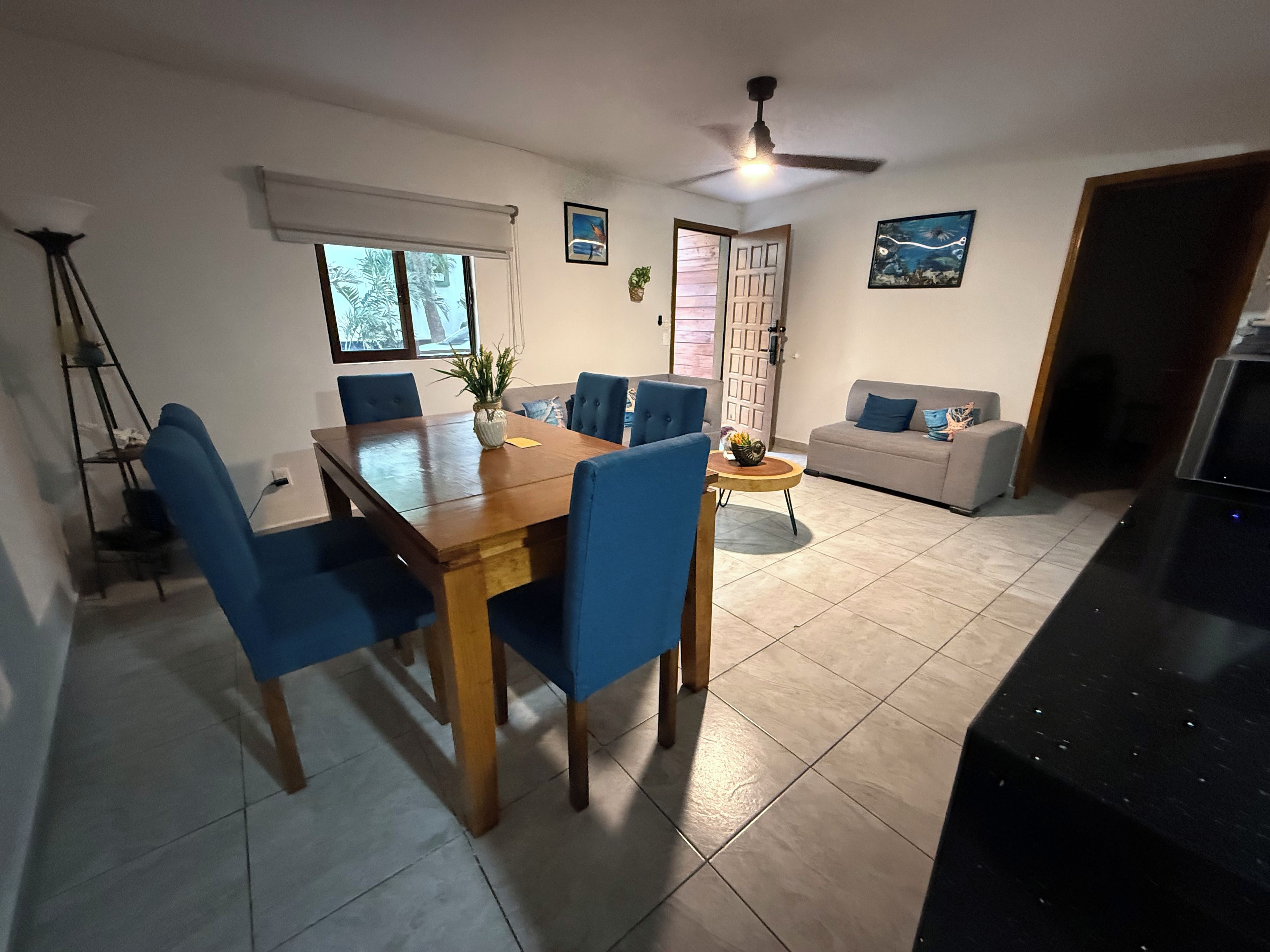 Ein Essbereich mit einem Holztisch, blauen Stühlen, einem grauen Sofa, einem Deckenventilator und einem Fernseher.
