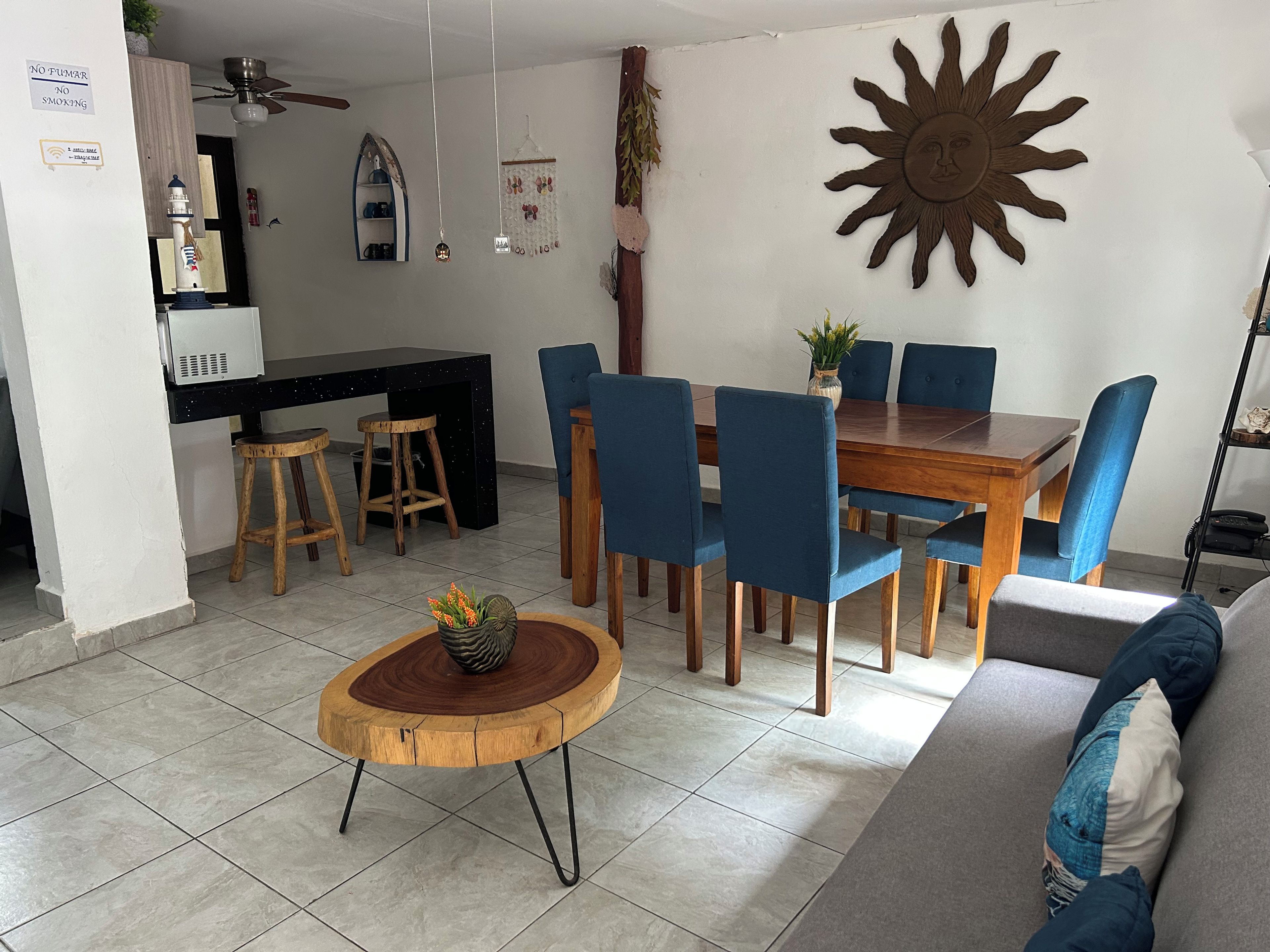 A dining area with a wooden table and blue chairs, a kitchen with a black countertop, and a sun-shaped wall decoration.