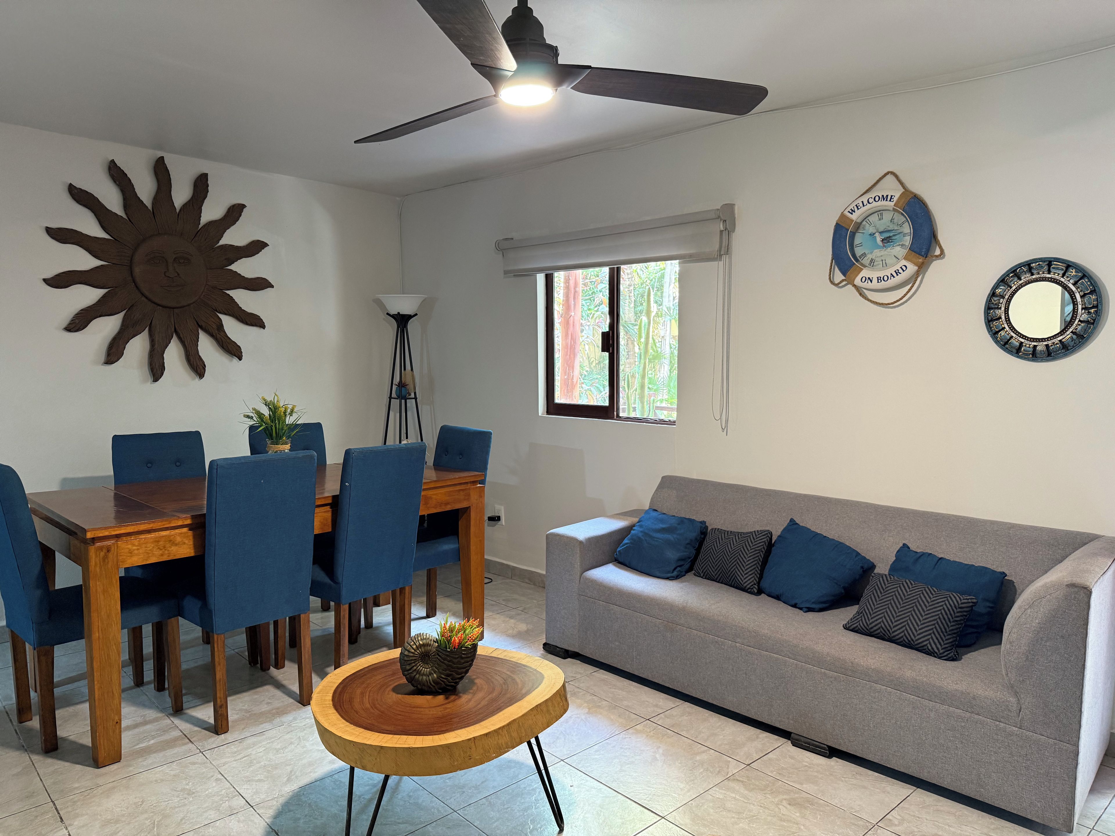 A dining area with a wooden table and blue chairs, a grey sofa with blue cushions, a decorative sun on the wall, and a ceiling fan.