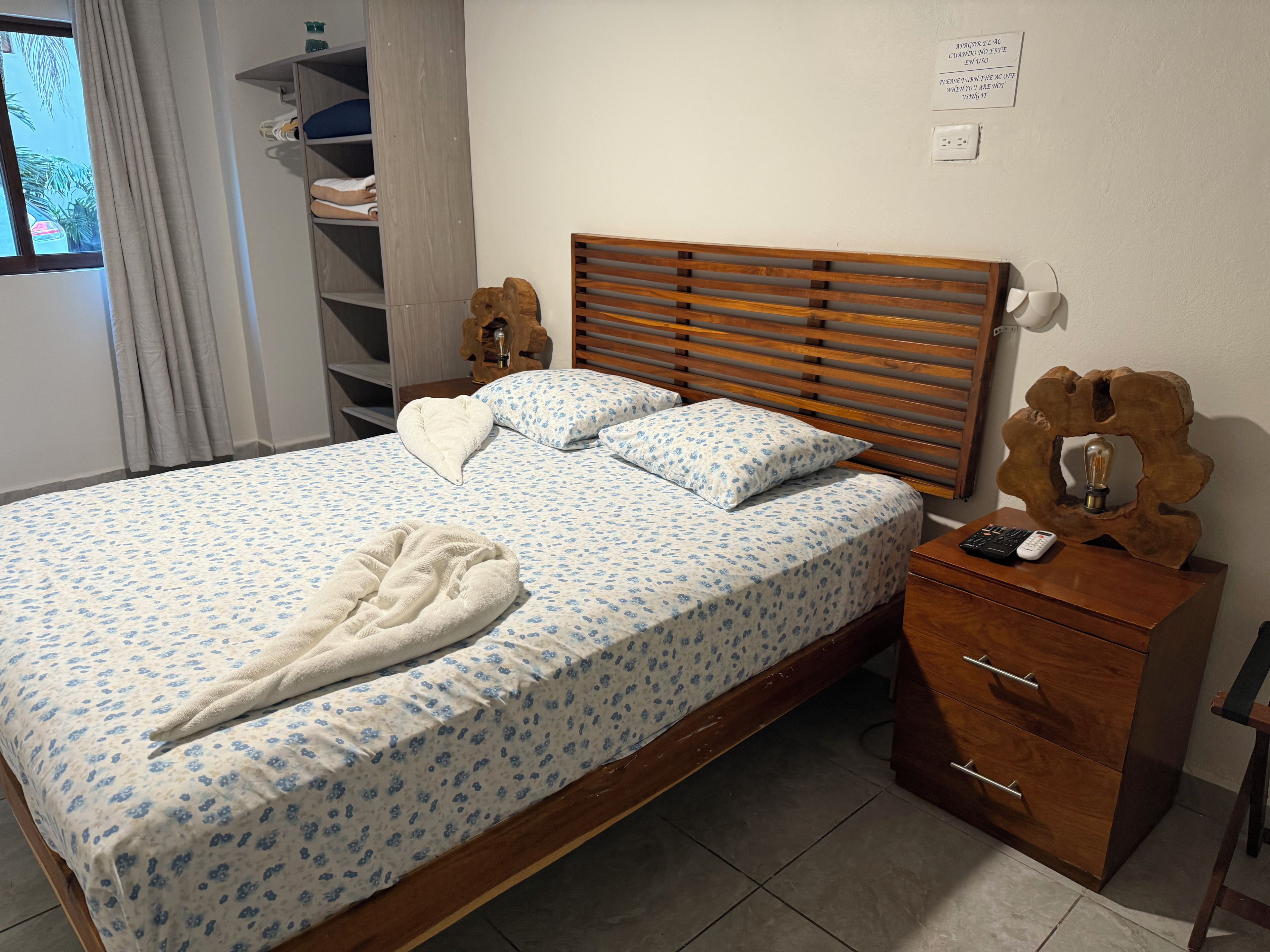Un lit bien fait, avec une couvre-lit à motifs floraux, une tête de lit en bois et une table de chevet sur laquelle se trouvent une lampe et une télécommande.