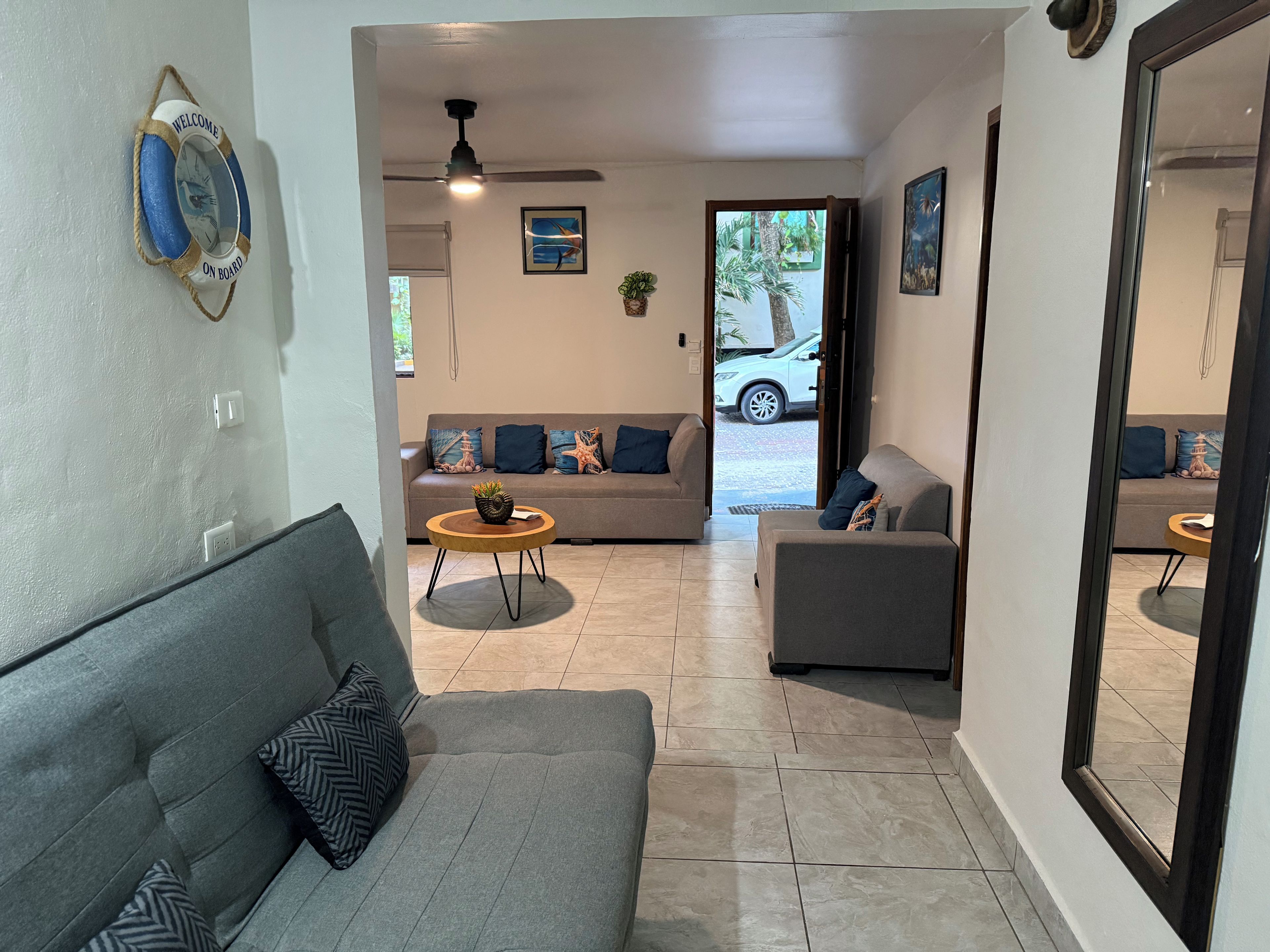 A living room with a grey sofa, a round wooden coffee table, and a grey armchair. There is a ceiling fan, a wall-mounted light fixture, and a framed picture on the wall.