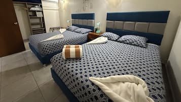A bedroom with two beds, each with patterned bedding and headboards. There is a nightstand with a lamp and a small plant on one bed.