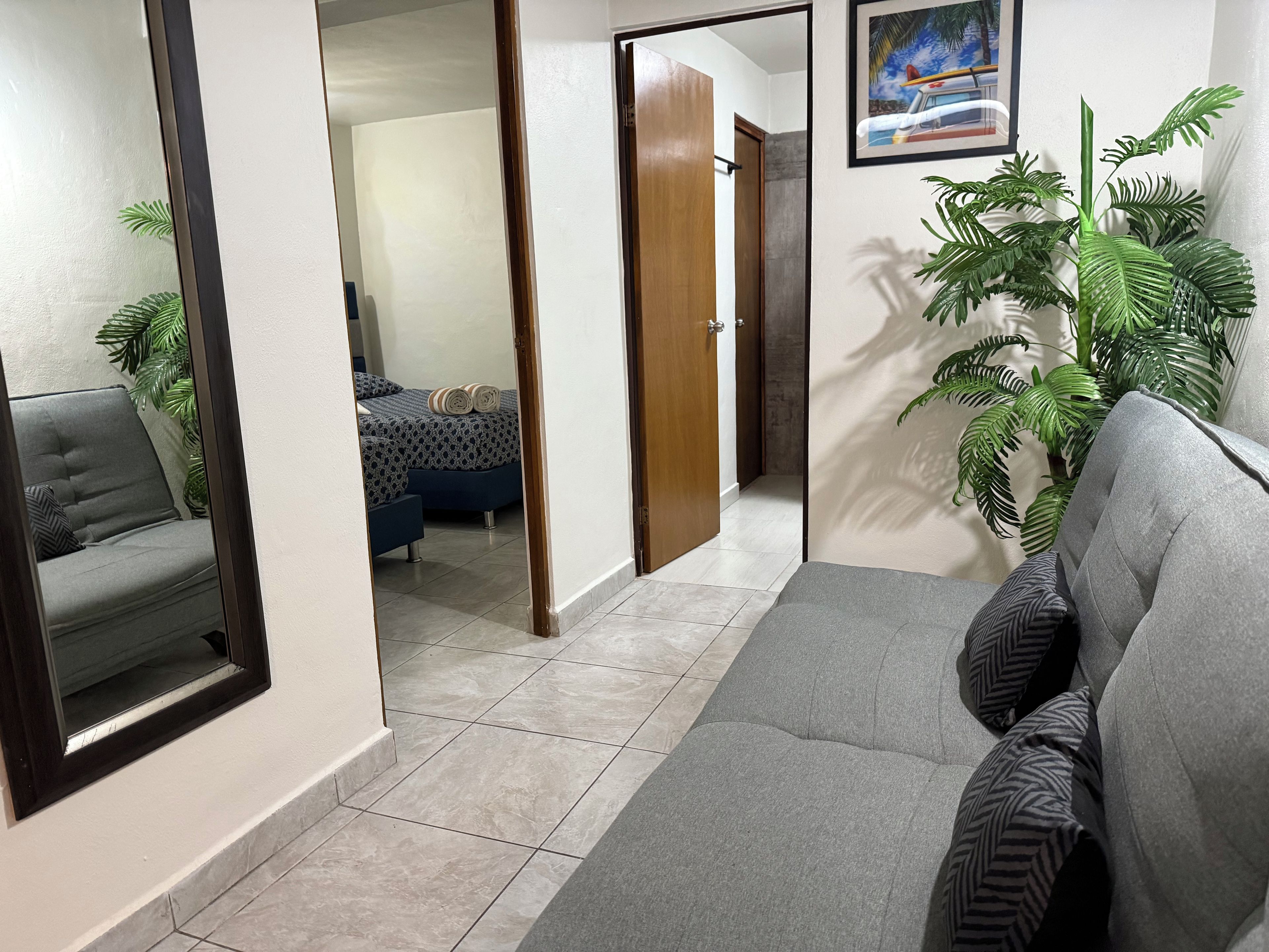 Un couloir avec un canapé gris, un miroir, un lit avec une literie à motifs et une plante en pot.