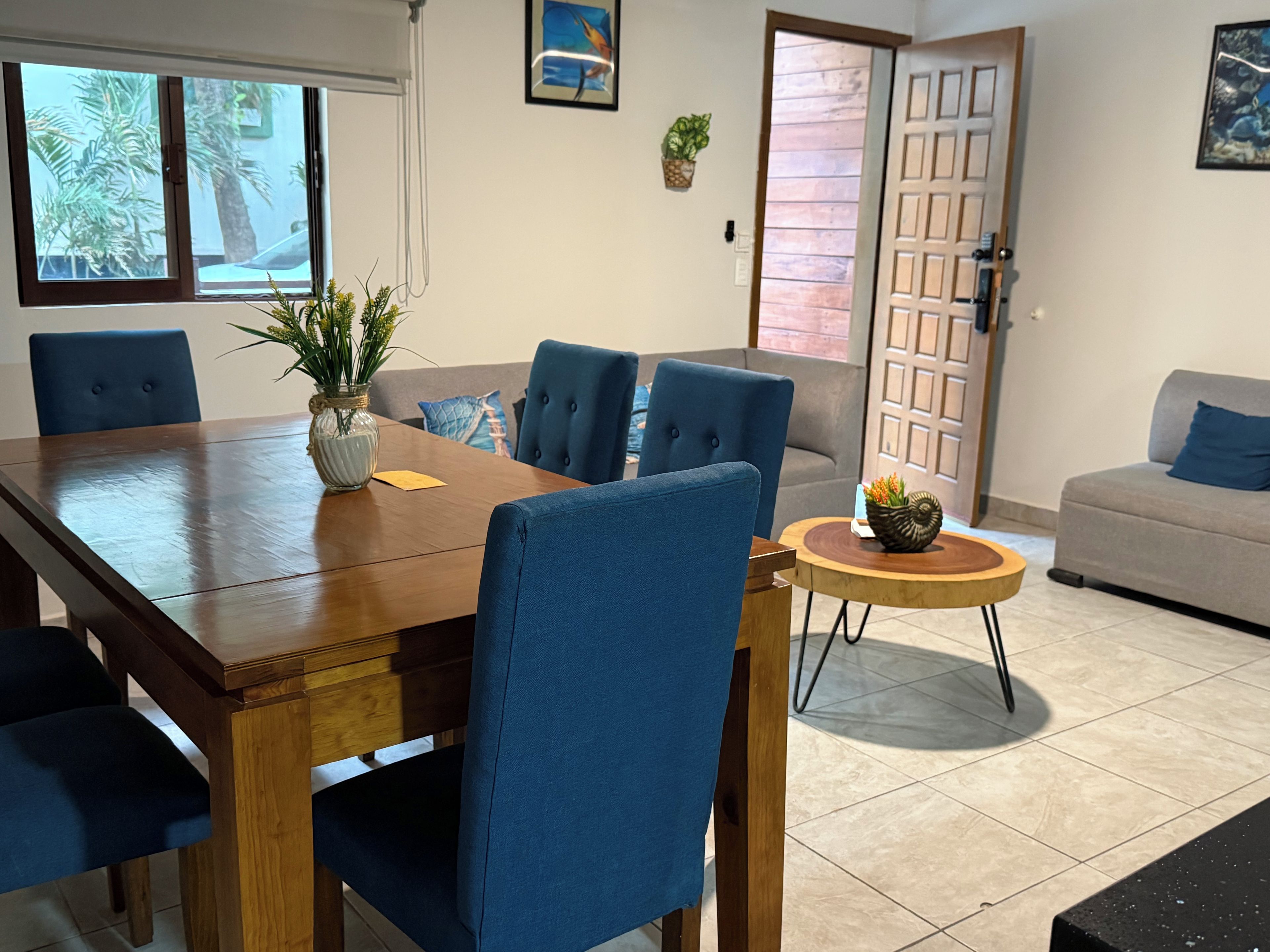 A dining area with a wooden table, blue upholstered chairs, a small round table with a plant, and a grey sofa in the background.