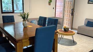 A dining area with a wooden table, blue upholstered chairs, a small round table with a plant, and a grey sofa in the background.