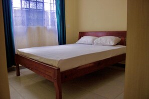 Una cama individual con estructura de madera y ropa de cama blanca en una habitación con piso de baldosas y ventana con cortinas.