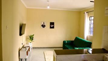 Una sala de estar con un sofá verde, un televisor de pantalla plana montado en la pared y un ventanal con cortinas.
