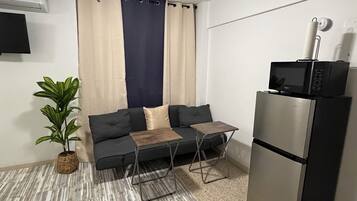 Un espace de vie compact comprenant un canapé, deux tables d’appoint, une plante en pot, un ventilateur de plafond, un four à micro-ondes et un réfrigérateur.