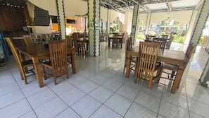 A covered outdoor dining area with wooden tables and chairs, a tiled floor, and a view of greenery in the background.