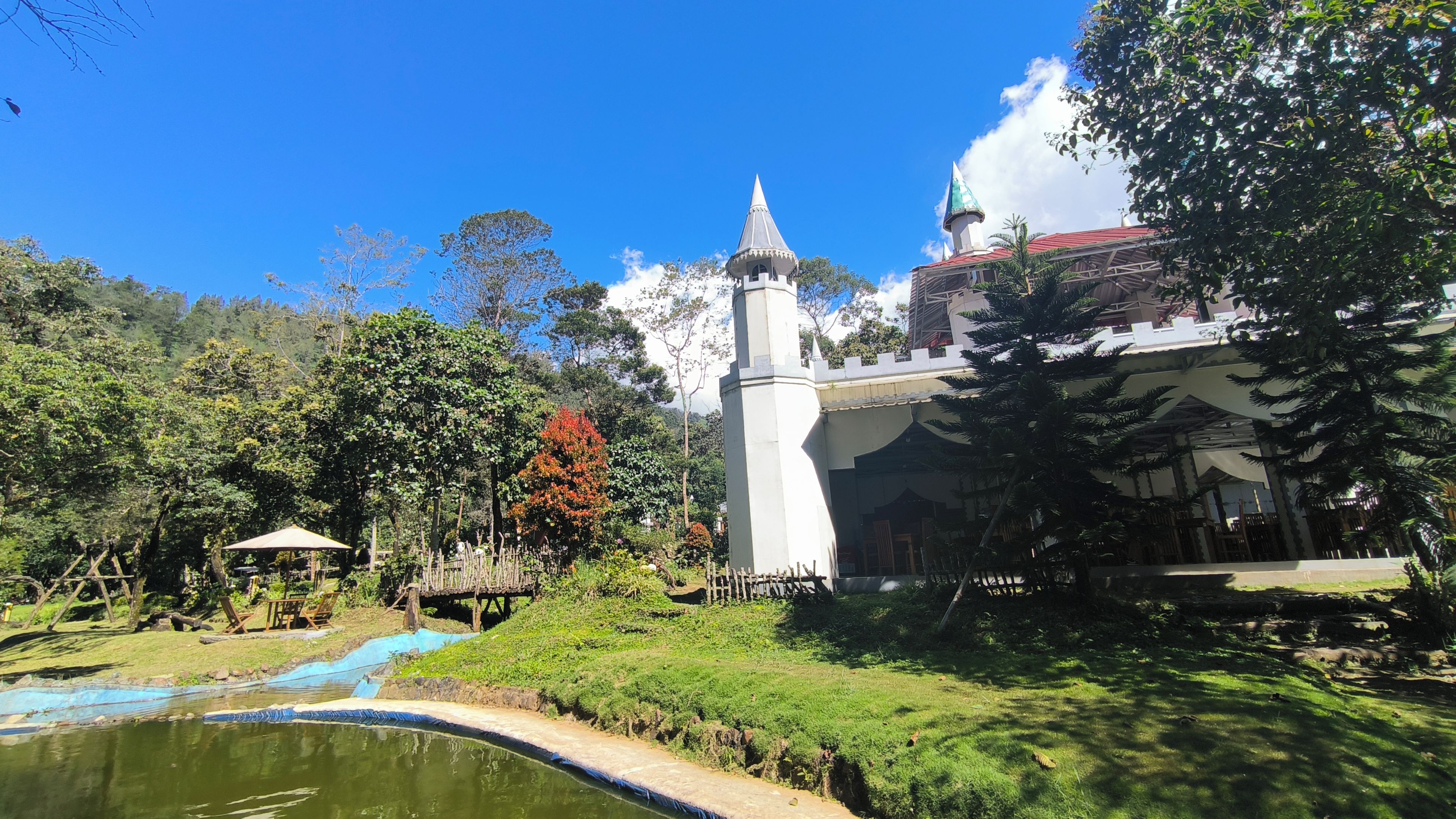 Un edificio parecido a un castillo con una torre, rodeado de exuberante vegetación y un pequeño estanque.