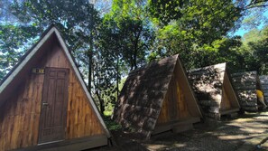 A row of wooden triangular huts with a door and a small porch area, surrounded by trees.