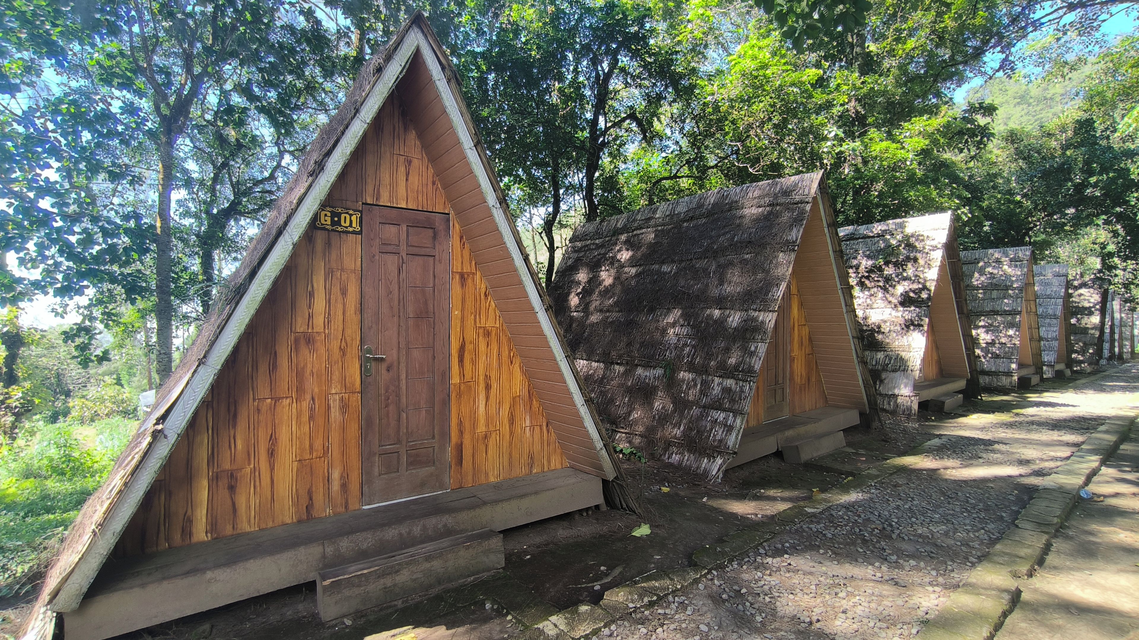 A row of wooden A-frame cabins with a door and a sign, surrounded by trees.