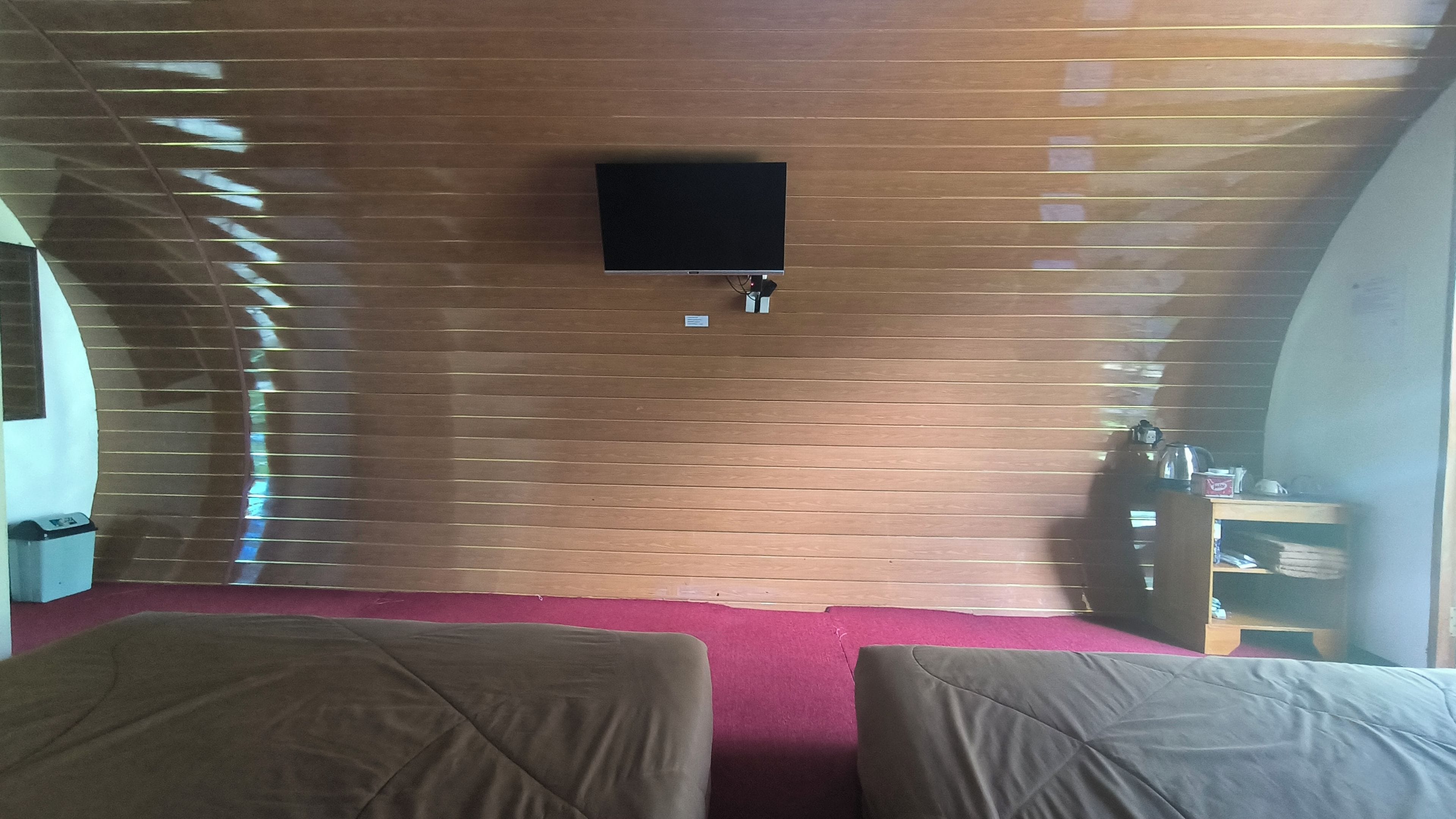A room with a flat-screen TV mounted on the wall, a small table with a kettle, and two beds with covers.