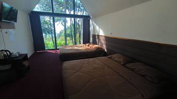 A hotel room with two beds, a TV mounted on the wall, and a large window with a view of greenery.