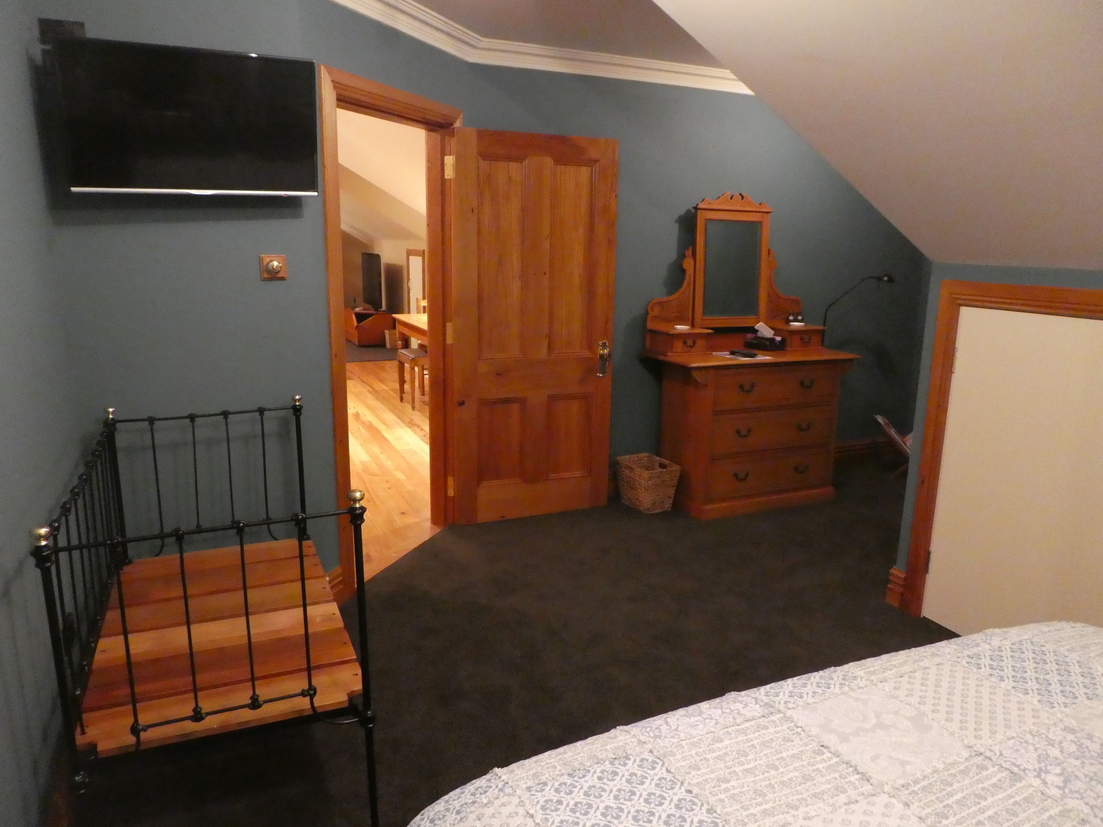 A bedroom with a bed, a wooden dresser, a mirror, a television on the wall, and a door leading to another room.