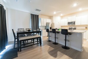 A modern kitchen with white cabinets, a central island with bar stools, and a dining area with a table and chairs. - Modern Oasis (Charlotte)