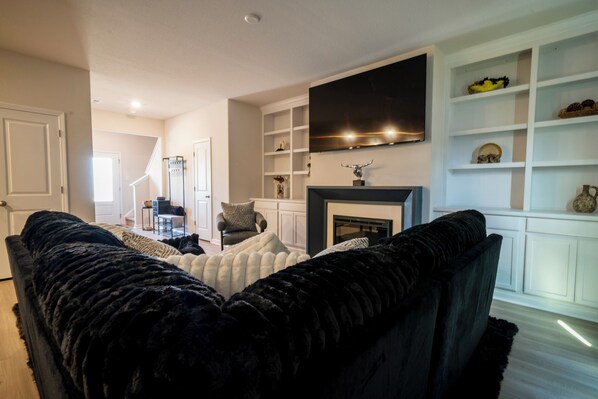 A living room with a black sofa, a fireplace, a television mounted above it, and a white shelving unit with decorative items. - Modern Oasis (Charlotte)