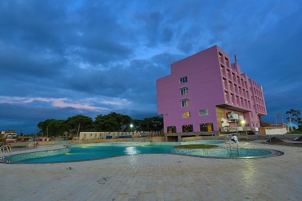 Un edificio moderno con zona de piscina, que incluye una piscina rectangular grande y una piscina circular más pequeña, rodeadas por un solárium con baldosas.