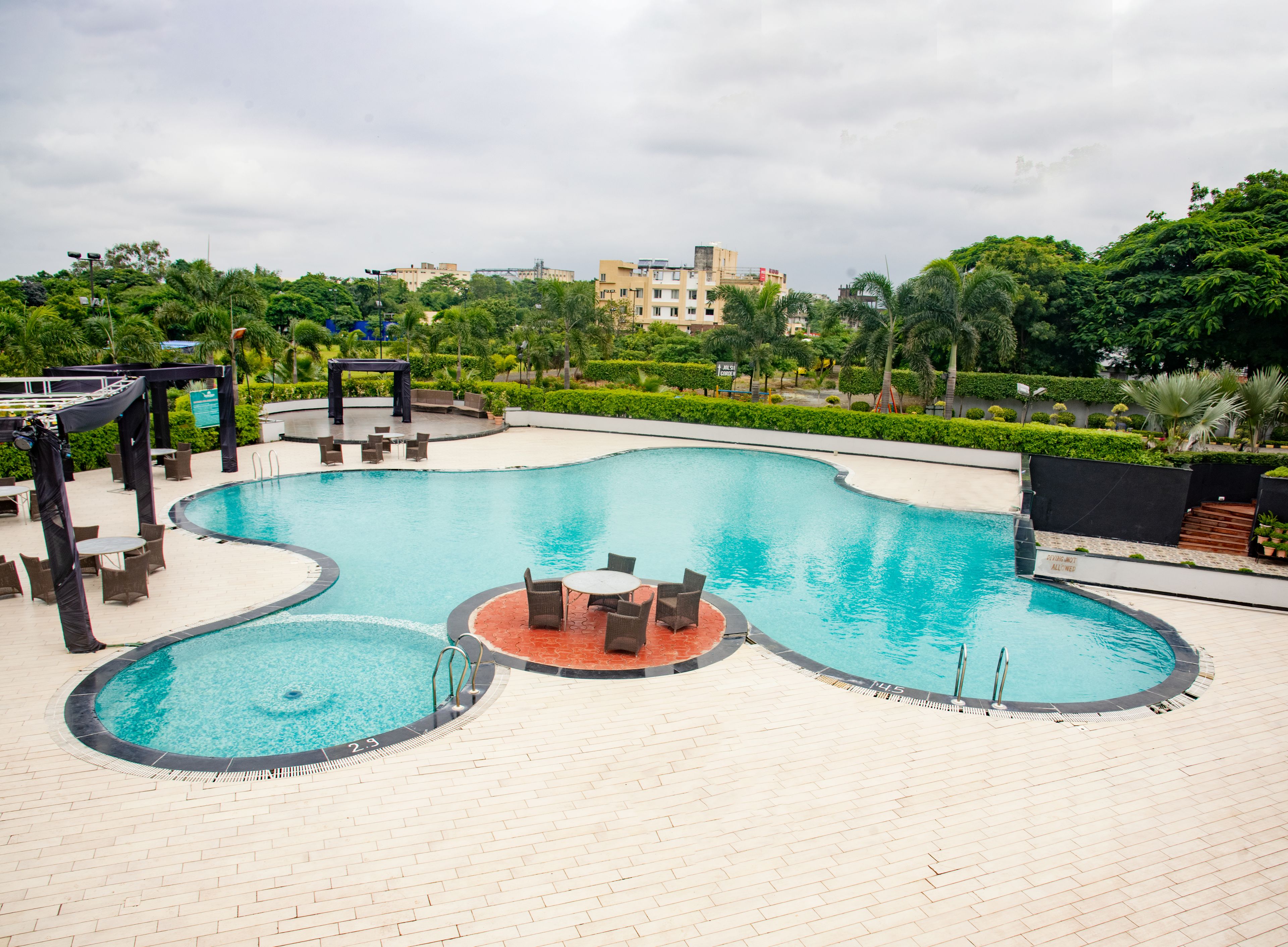A swimming pool with a curved design, surrounded by lounge chairs and tables, and a backdrop of greenery and buildings.