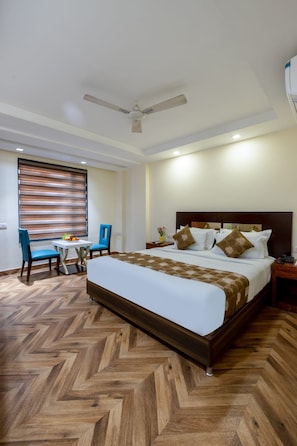 Una camera d'albergo con un letto grande, una ventola a soffitto, un tavolino con sedie e una finestra con persiane.