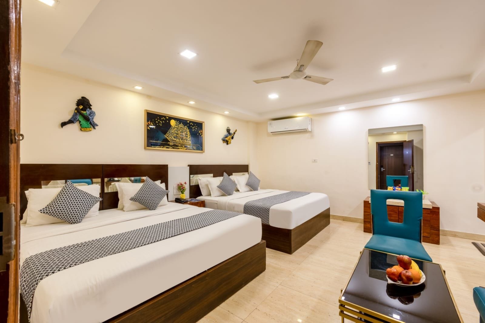 A hotel room with two beds, a ceiling fan, a painting, and a fruit bowl on a table.