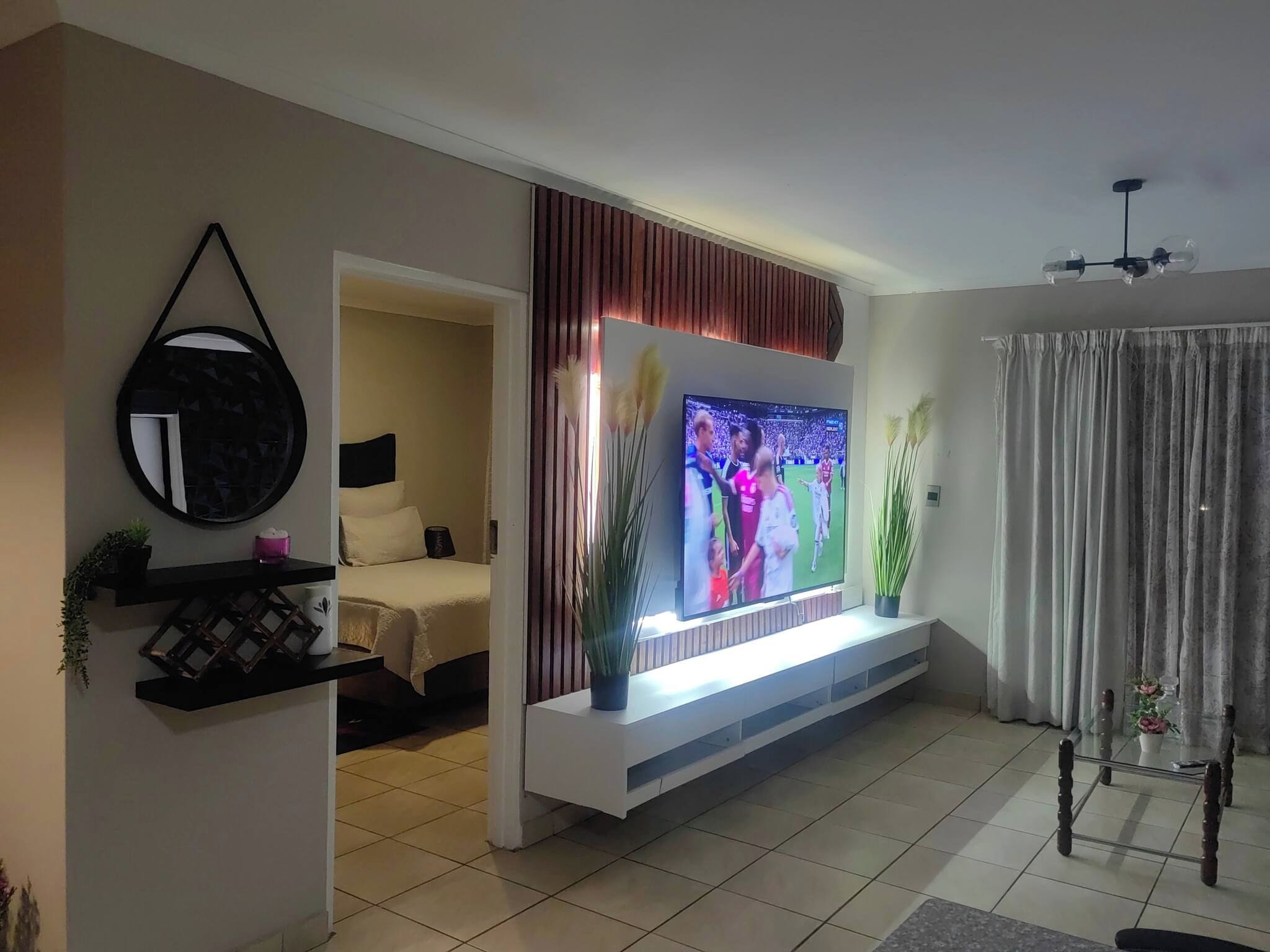 A living room with a flat-screen TV mounted on a white console, a bed with a headboard, a mirror, and a potted plant.