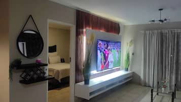 A living room with a flat-screen TV mounted on a white console, a bed with a headboard, a mirror, and a potted plant.