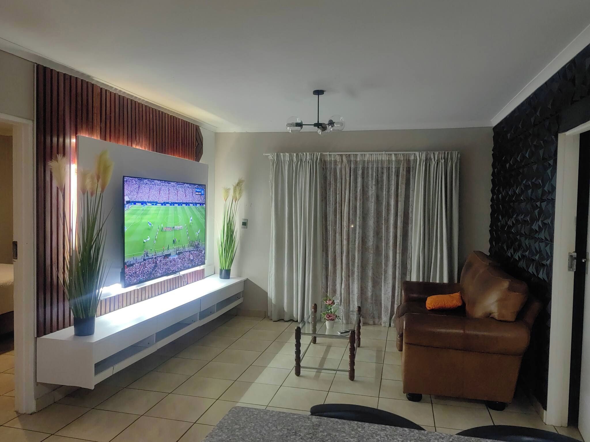 A living room with a flat-screen TV mounted on a wall, a brown leather sofa, and a large window with curtains.