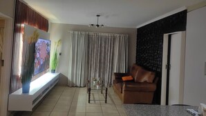 Uma sala de estar com sofá, mesa de centro, televisão e janela com cortinas.