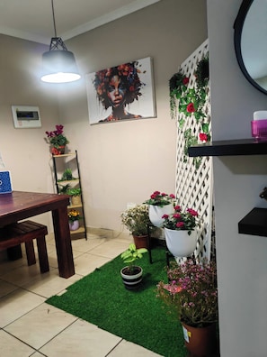 A dining area with a wooden table, chairs, a hanging light fixture, a wall-mounted thermostat, and a potted plant.